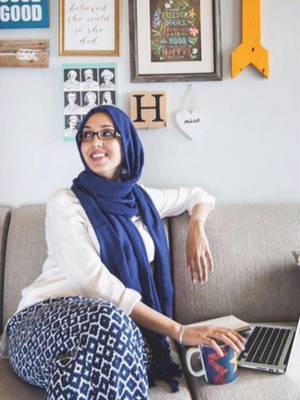 a lady in hijab looking away while sitting on a sofa with her laptop and hand on a mug